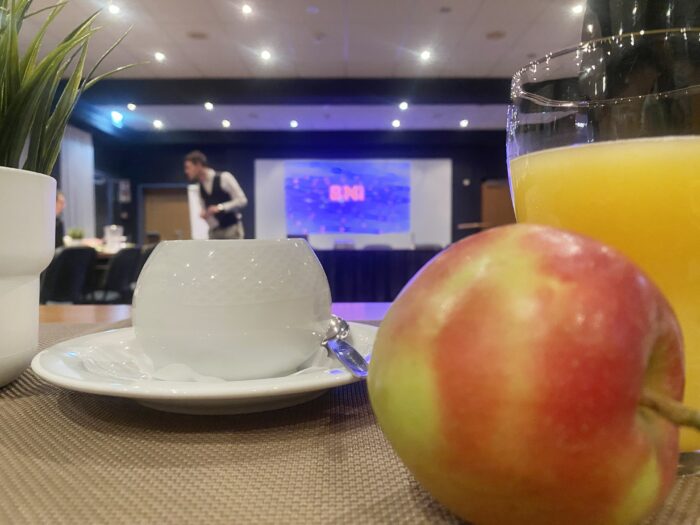 Ein Konferenzraum mit gedimmter Beleuchtung. Im Vordergrund steht ein Getränkeglas mit Orangensaft, ein roter Apfel und eine leere, weiße Kaffeetasse auf einem Unterteller, daneben ein kleiner Topf mit einer grünen Pflanze. Im Hintergrund bereitet ein Mann in einem Anzug den Raum vor, während auf einem unscharfen Bildschirm Präsentationsfolien zu sehen sind. Der Raum ist für ein professionelles Event vorbereitet.